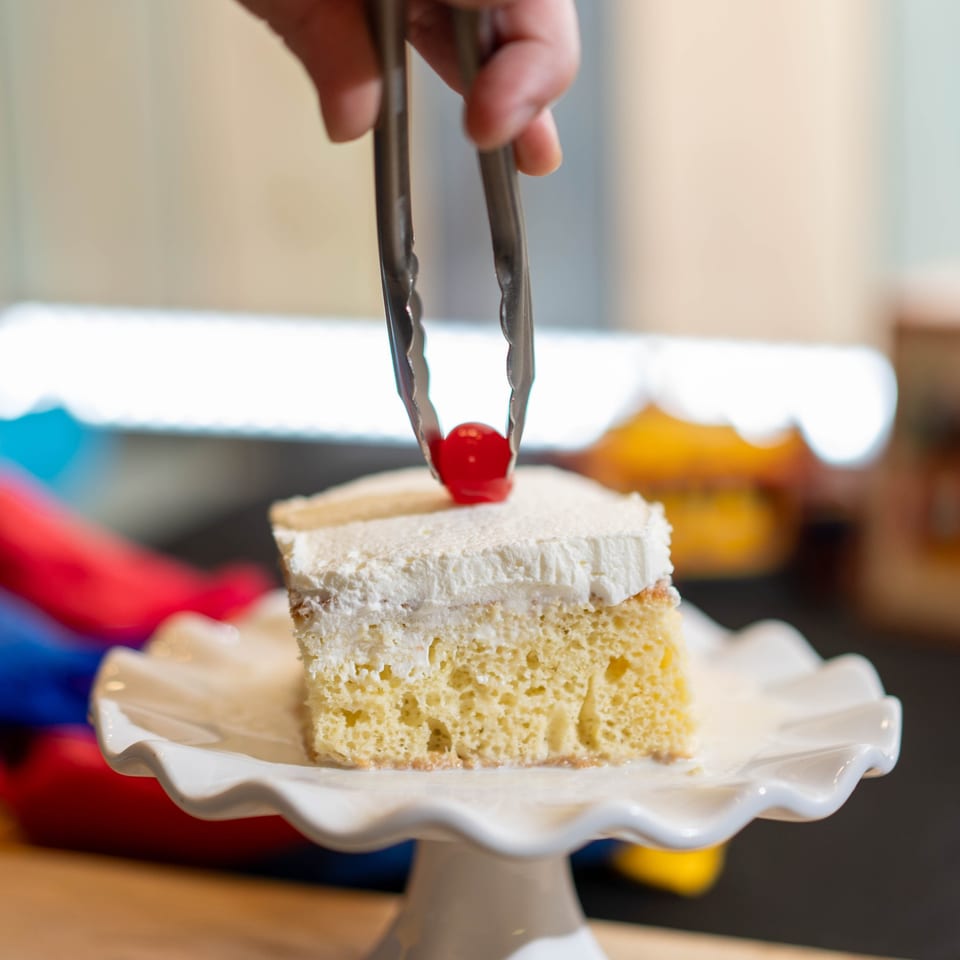 A hand uses tongs to place a red cherry on a slice of tres leches cake with whipped cream.