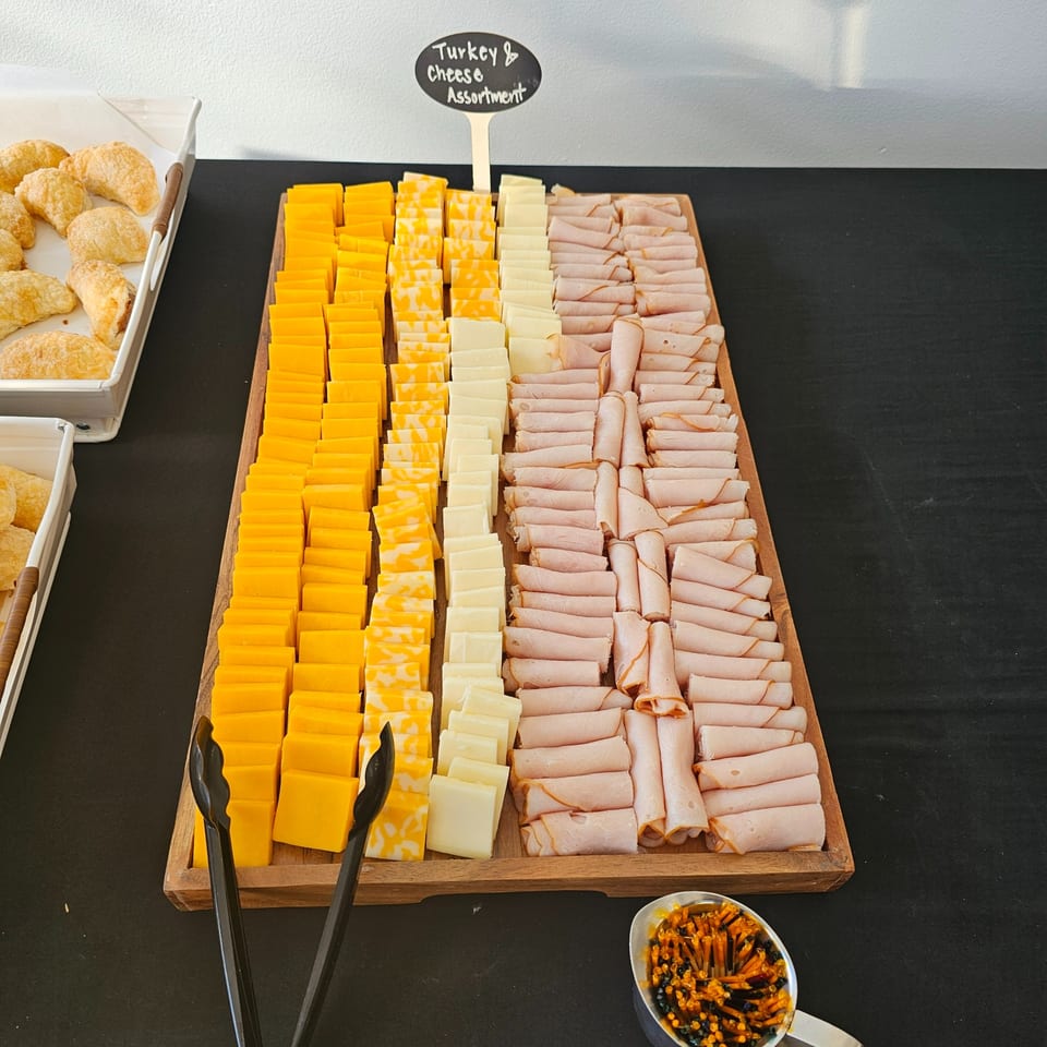 A wooden tray holds rows of sliced turkey, cheddar, and marble cheeses, with a sign reading "Turkey & Cheese Assortment."