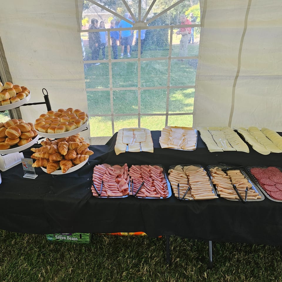 A buffet table under a tent with trays of bread, croissants, sliced meats, and cheeses.