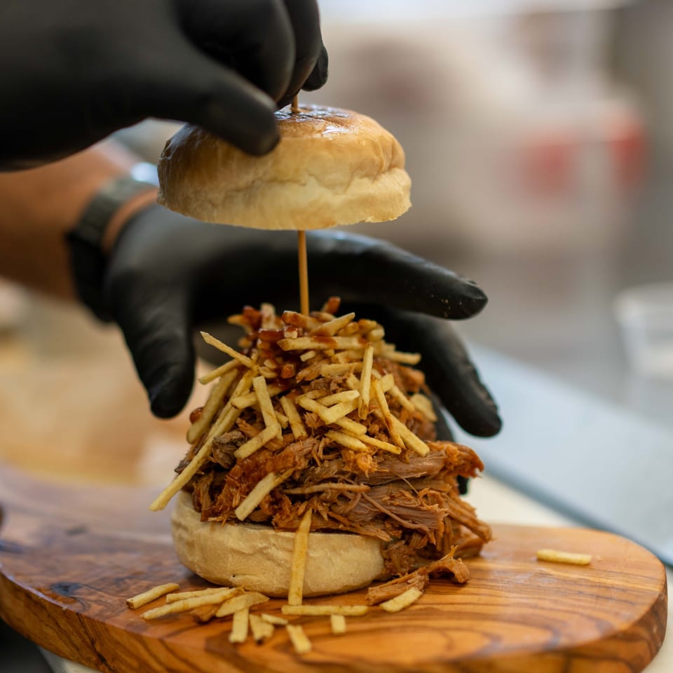 A chef's gloved hands assemble a La Boca Loca burger with pulled pork and crispy fries.