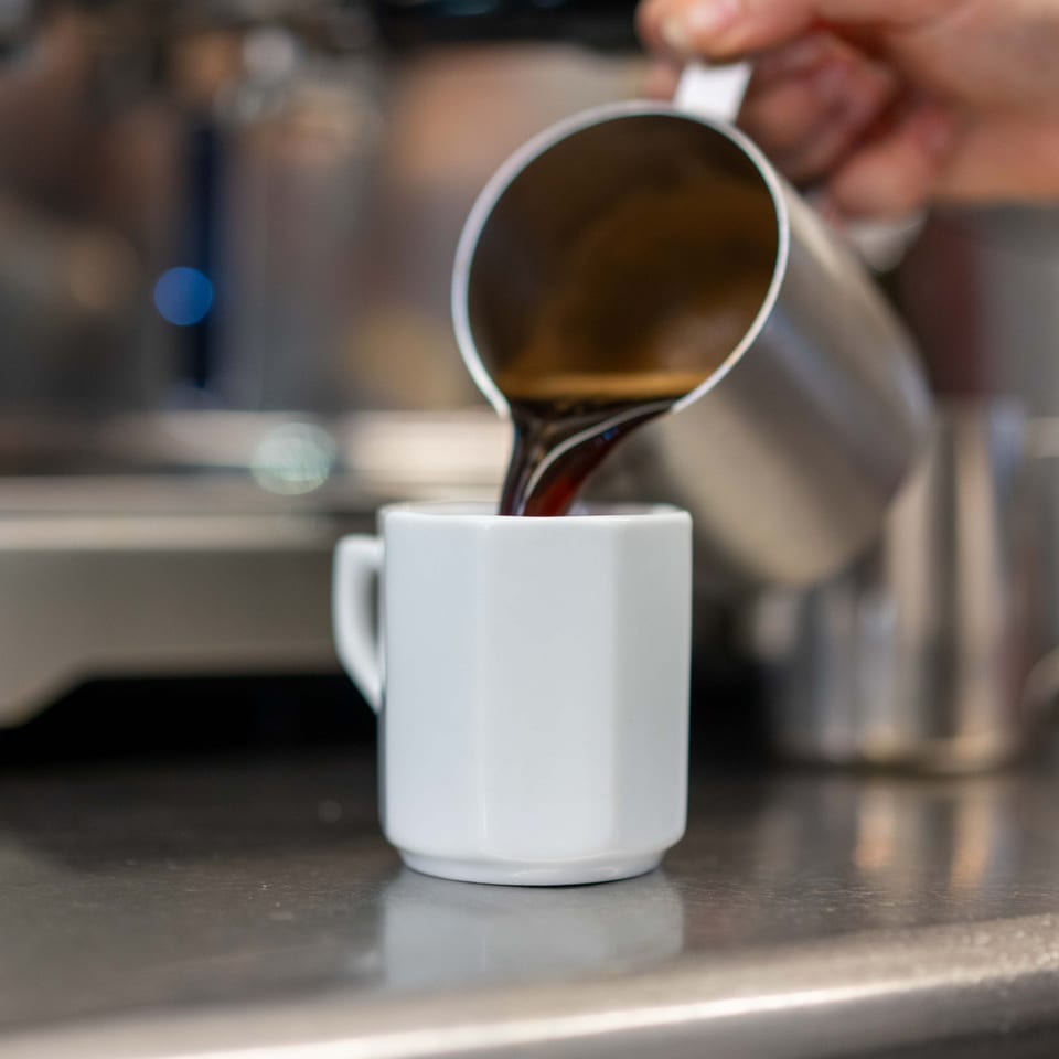 A hand pours dark coffee from a silver pitcher into a small white cup.
