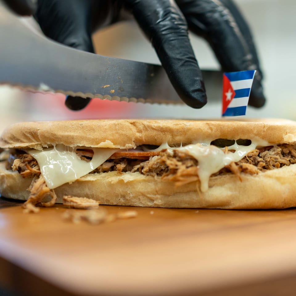 A hand in a black glove cuts a Cuban sandwich with melted cheese and shredded meat.