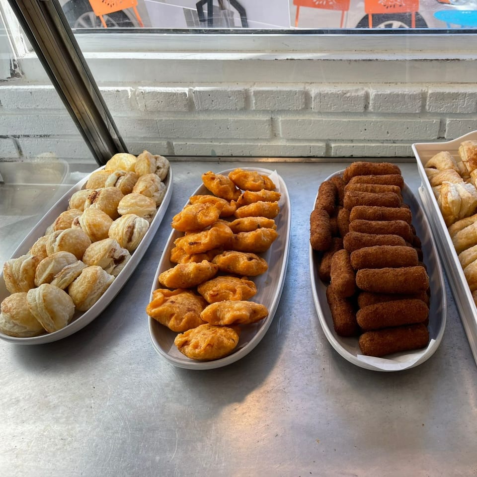 Three white trays filled with various pastries and croquettes on a metal counter by a window.